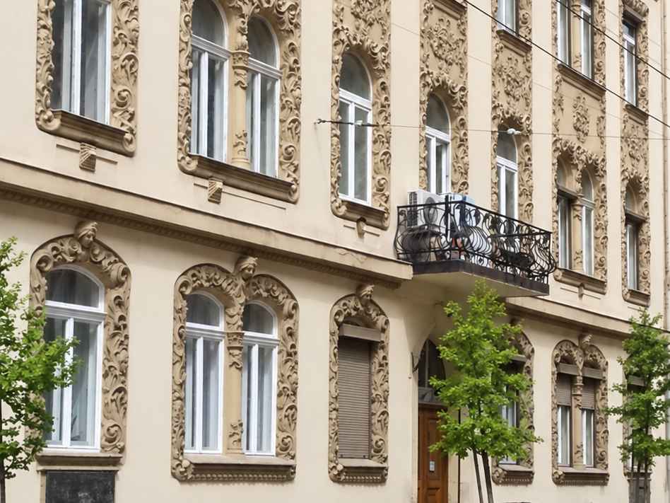 A Dembinszky utca 18. földszintje és első emelete ferde rálátásból: arcos zárókövű, dúsan faragott ablakkeretek, kovácsoltvas erkély, a kapu előtt fiatal utcafák
